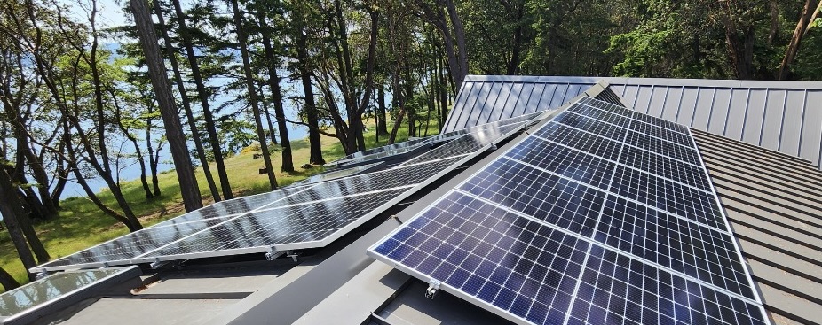 a roof-top view of a solar panel installation on top of a metal roof. The home is beside many trees and the ocean is visible through the tree line.