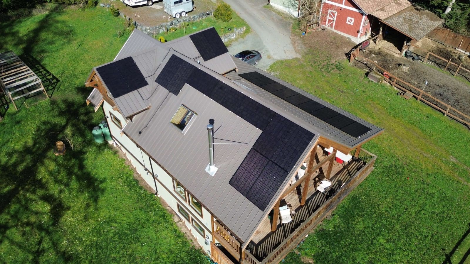 An aerial view of a home with multiple arrays of solar panels on the roof. The home is in a rural setting with farm buildings and a horse stable to in the background.